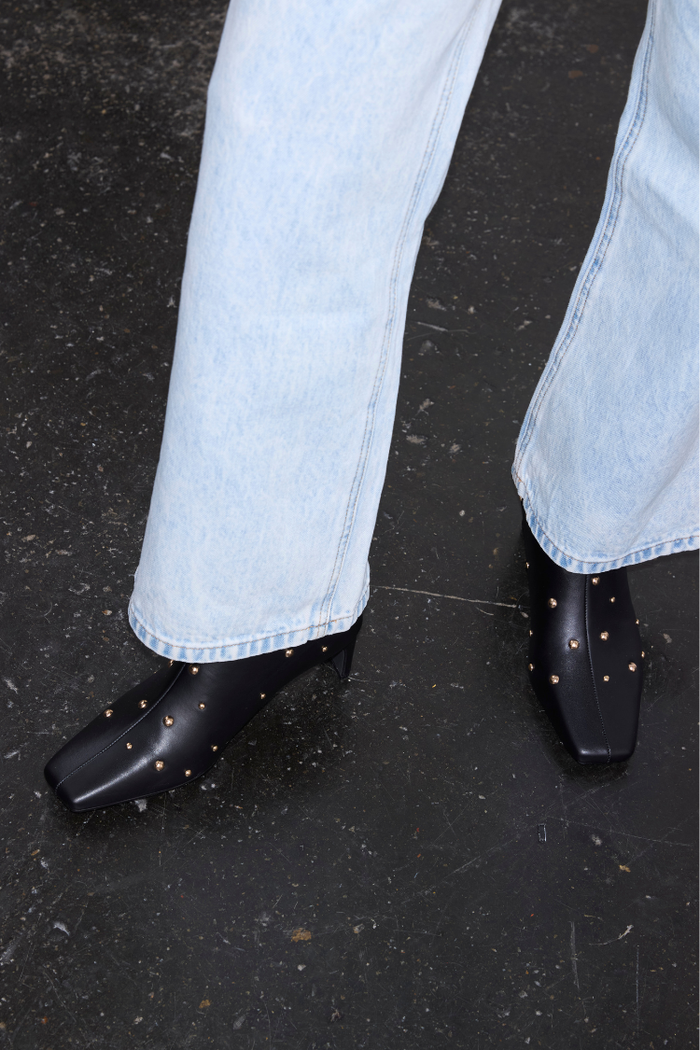 Black leather ankle boots with gold metal studs and a square toe, paired with light-wash denim jeans.