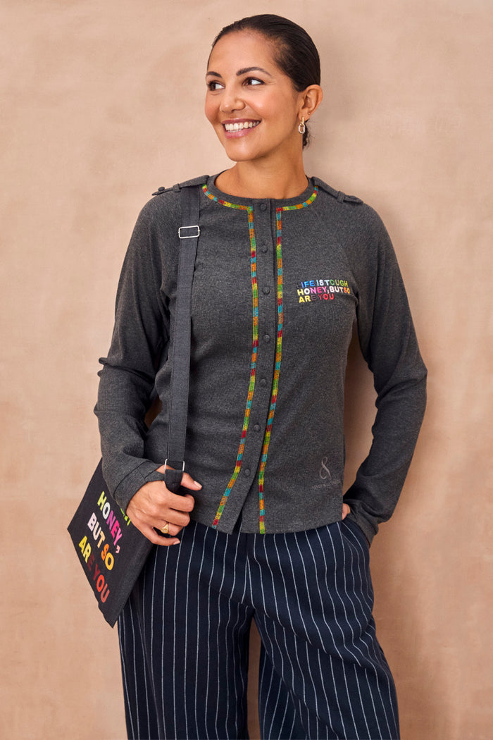 Charcoal Life is Tough Honey drainage bag with multi-coloured text, styled with a matching top and pinstripe trousers.