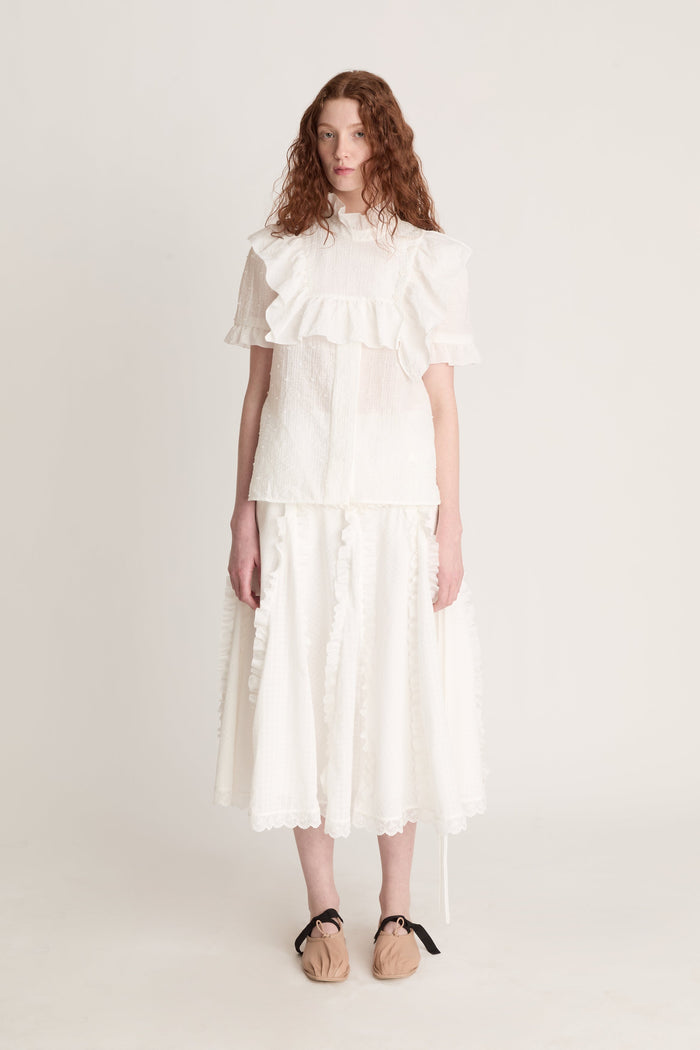 A woman wearing a white top with ruffled sleeves  and a white skirt on a plain background