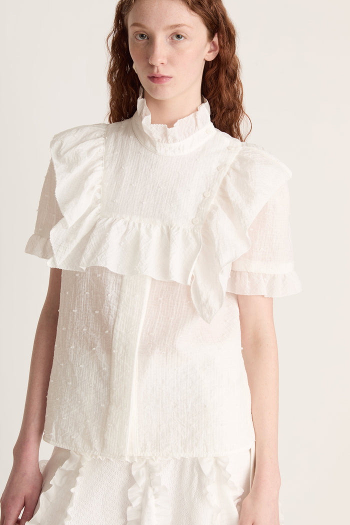 A woman wearing a white top with ruffled sleeves  and a white skirt on a plain background
