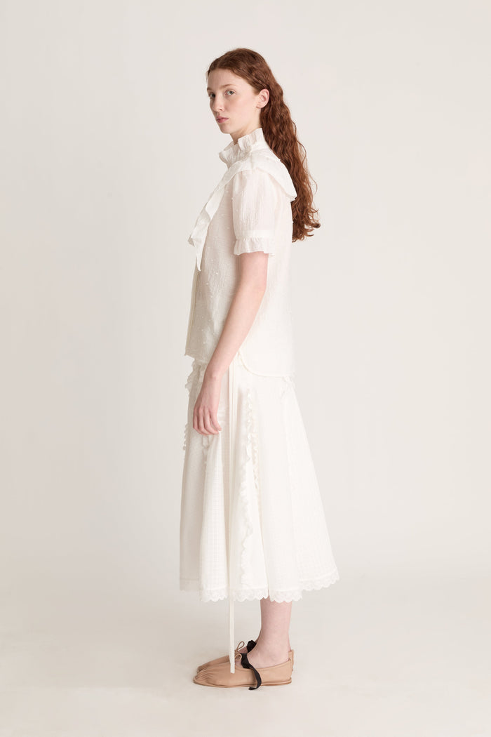 A woman wearing a white top with ruffled sleeves  and a white skirt on a plain background